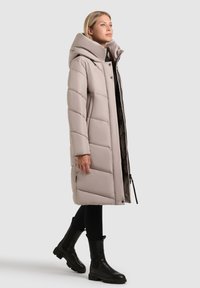 Kvinde iført en lang beige pufjakke med høj krave, sorte leggings og sorte ankelstøvler, stående mod en ensfarvet lys baggrund.