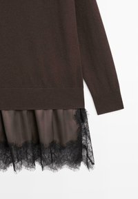 Pull à manches longues marron foncé superposé sur une jupe marron avec une bordure en dentelle noire à l'ourlet, sur fond blanc.