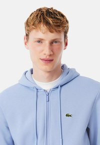 Hoodie zippé bleu clair en tissu doux, avec une poche kangourou et un logo brodé vert sur la poitrine gauche.