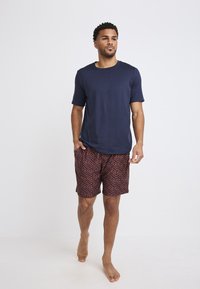 Marineblauwe crew-neck T-shirt gecombineerd met gestreepte shorts met een rood-blauw zigzagpatroon. Blootsvoets, met handen in de zakken.