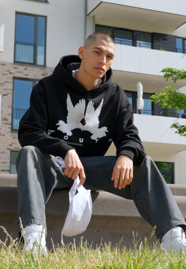 Black hooded sweatshirt with white bird graphic, paired with loose grey jeans and white sneakers. Sitting on a stone bench in a grassy area.