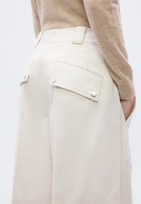 Φαρδιά παντελόνια σε ανοιχτό μπεζ χρώμα με δύο πίσω τσέπες που κλείνουν με κουμπιά, με λεία υφή και ραμμένη μέση.