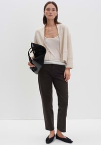 Beige verkürzter Cardigan über einem weißen Tanktop, kombiniert mit dunklen braunen Hosen und schwarzen flachen Schuhen. Das Modell hält eine schwarze Netz-Tasche.