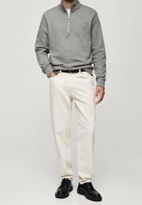 Sudadera gris con cremallera, pantalones blancos de pierna recta y zapatos negros con cordones. La sudadera tiene un cuello alto y puños acanalados.