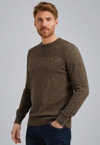 Pull en texture marron avec un col et des épaules côtelés, arborant un petit logo sur la poitrine. Porté avec un jeans foncé.