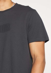 Czarna bawełniana koszulka z okrągłym dekoltem, krótkimi rękawami i subtelnym wytłoczonym logo z przodu, pokazana na jednolitym tle.