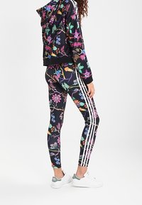 Svart huvjacka och leggings i tyg, prydda med färgglada blommönster. Tre vita ränder löper längs leggings och ärmar på jackan.