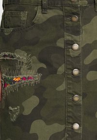 Camouflage spijkerrok met knoopsluiting aan de voorkant, kenmerken van versleten stukken die de bloemmotiefstof eronder onthullen en contrasterende stiksels.