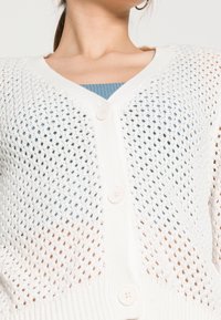 Cardigan tricoté blanc avec un col en V, présentant un tissage lâche et ouvert et trois boutons sur le devant. Motif texturé sur l'ensemble.