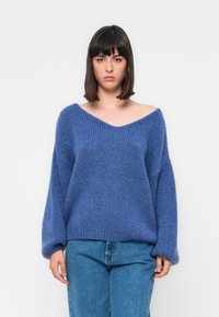 Maglione blu oversize a maglia con scollo a V, maniche lunghe e tessuto texture. Abbinato a jeans blu chiaro, su uno sfondo semplice.