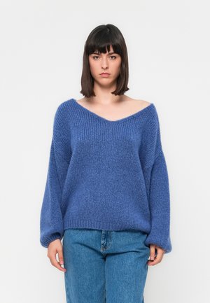 Übergroßer blauer Strickpullover mit V-Ausschnitt, langen Ärmeln und texturiertem Stoff. Kombiniert mit hellblauen Jeans, vor einem schlichten Hintergrund.