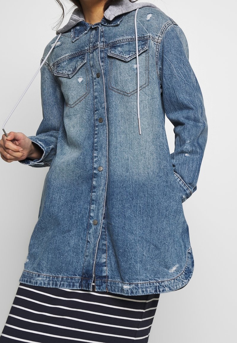 Veste en jean avec capuche grise, fermeture à boutons, deux poches poitrine et détails usés. Manches longues et ourlet arrondi avec couture contrastante.