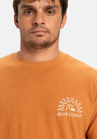 Oranžové bavlněné tričko s kulatým výstřihem a bílým logem se sluncem a vlnou na levé části hrudi.