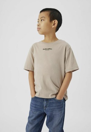 Beige bomulls t-shirt med korta ärmar och rund halsringning, med svart text som säger "studio edition" på bröstet. Bärs med blå jeans.