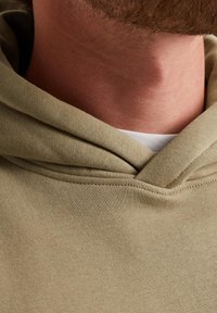 Beige Baumwoll-Hoodie mit einer doppelten Kapuze, sichtbaren Nähten und einem weißen Innenkragen, der eine Nahaufnahme des Kragens und der Stoffstruktur zeigt.