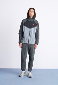 Track suit featuring a black and gray color scheme, with a zip front, stand-up collar, and subtle logo accents. Smooth fabric, fitted design.