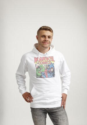 Junger Mann trägt weißen Marvel Comics Hoodie mit Superhelden-Grafiken und steht mit verschränkten Händen vor einfarbigem Hintergrund.
