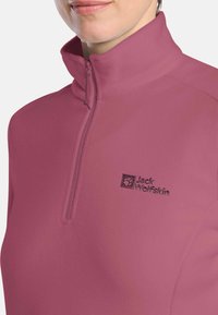 Primo piano di una persona che indossa una giacca in pile Jack Wolfskin di color mauve con zip a un quarto e colletto alto.