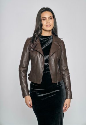 Vrouw met lang donker haar die een bruine leren jas draagt over een zwarte fluwelen jurk, staande tegen een effen lichtgrijze achtergrond.