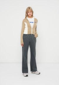 Beige og hvit jakke med glidelås og blå logo, kombinert med mørkegrå vide jeans og hvite sneakers. Glatt tekstur, avslappet passform.