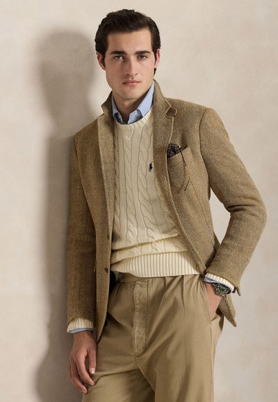 Homme portant un blazer beige en chevrons, un pull en maille torsadée crème, une chemise bleu clair, un pantalon beige et une montre, debout devant un mur neutre.