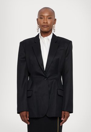 Blazer noir oversize avec épaules structurées, fermeture à un bouton et poches avant. Porté sur une chemise blanche.