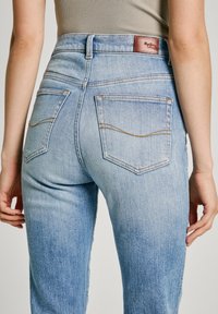 Ljusblå denimjeans med hög midja, med bakfickor och kontrasterande sömmar. Midjebandet har en brun läderlapp.