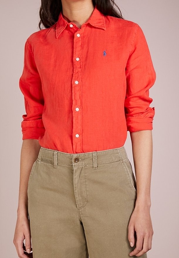 Red linen button-up shirt with rolled-up sleeves and a small blue logo on the left chest. Paired with beige cotton trousers.