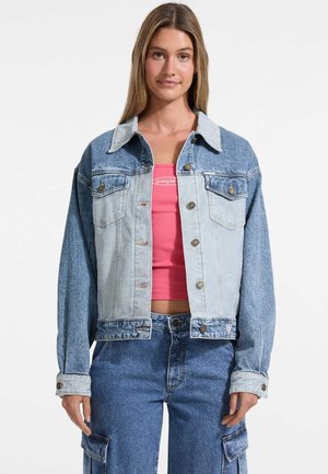 Veste en denim bleu clair avec un empiècement contrasté de bleu foncé. Elle possède des poches poitrine, des fermetures à boutons et une coupe décontractée. Portée sur un haut rose.