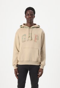 Béžová mikina s klokankou, so viacfarebným logom "GAP". Kapucňa má károvanú podšívku. Model má na sebe čierne džínsy.