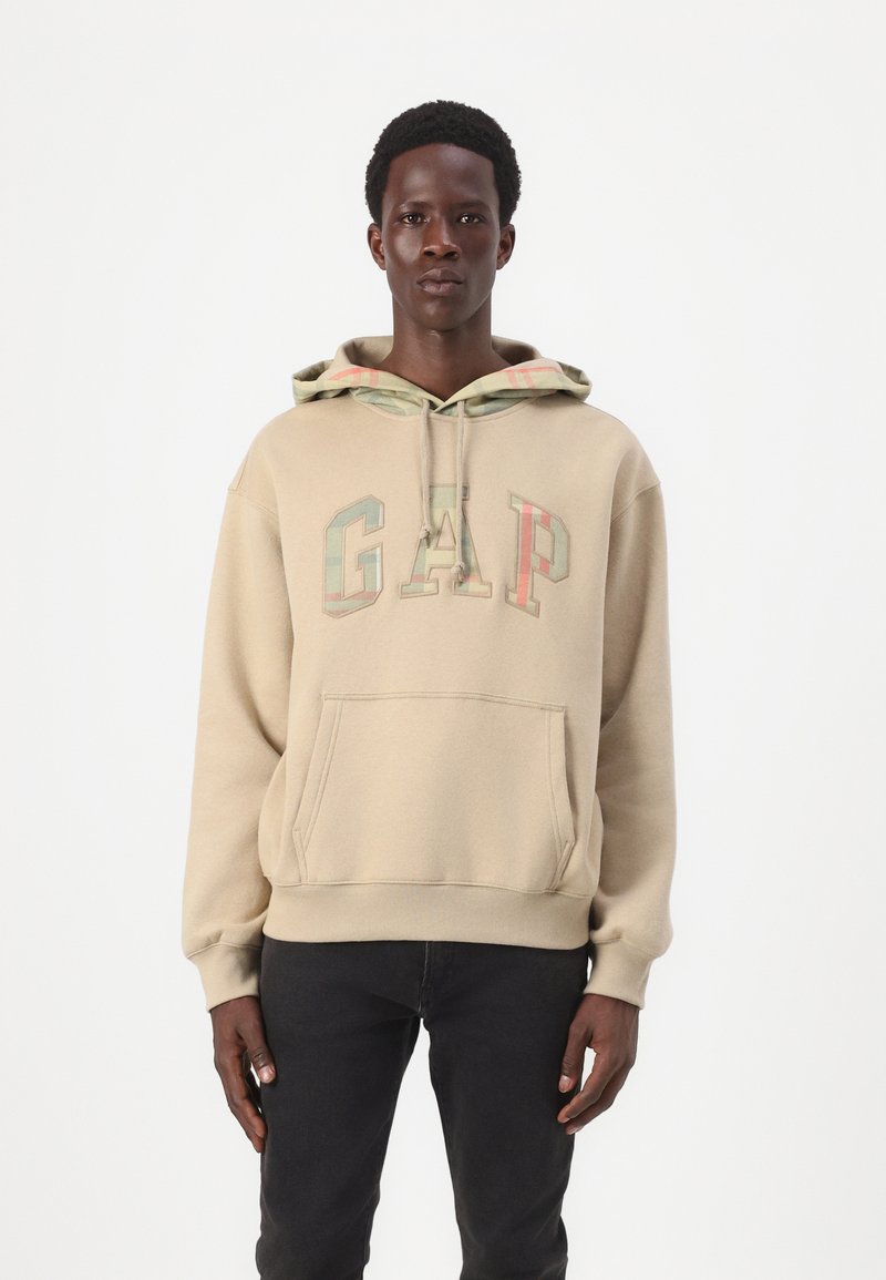 Béžová mikina s klokankou, so viacfarebným logom "GAP". Kapucňa má károvanú podšívku. Model má na sebe čierne džínsy.