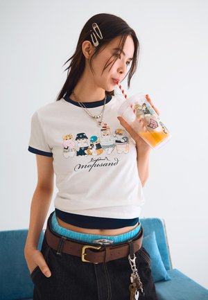 T-shirt bianco a maniche corte con bordi blu navy, caratterizzato da personaggi dei cartoni animati colorati. Jeans neri a vita alta con una cintura marrone e una tazza trasparente con una cannuccia.