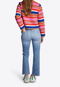 Femme portant un jean bleu clair avec des motifs de fleurs roses et un pull rayé coloré, se tenant avec une main dans la poche.
