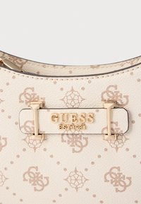 Kermansävyinen laukku, jossa hienovarainen beige logokuvio. Kultaiset metalliosat, joissa suorakaiteen muotoinen laatta, johon on kaiverrettu "GUESS EST. 1981".