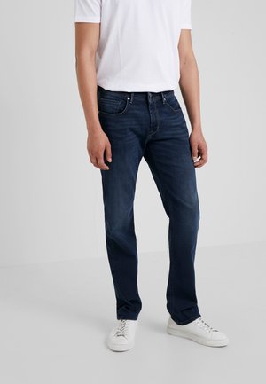 Homme portant un jean bleu foncé, un t-shirt blanc et des baskets blanches, debout devant un fond gris uni.