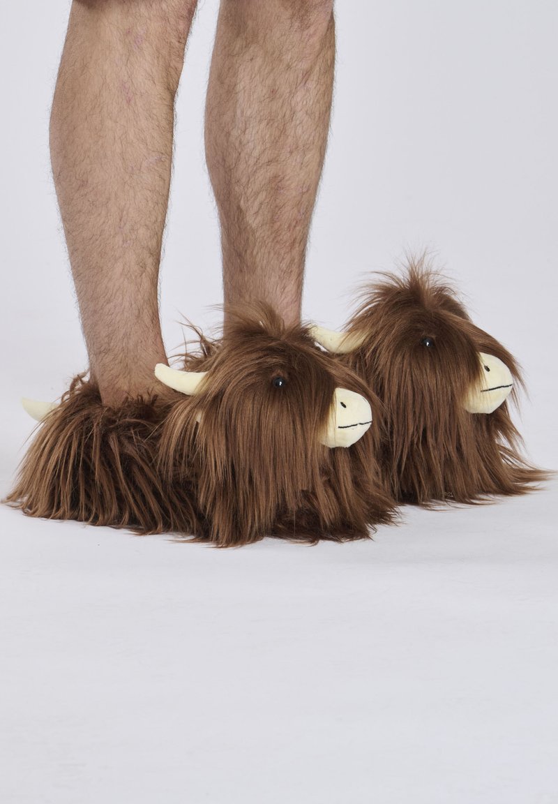 Braune, fuzzy Hausschuhe in Form von Tierköpfen mit Hörnern, die eine lange, haareähnliche Textur aufweisen und schwarze Knopfaugen haben. Beinen sind teilweise sichtbar.