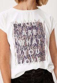 Bílá kratší košile s krátkým rukávem, na přední straně s barevným abstraktním čtvercem a textem "JSTE TO, CO MILUJETE", nošena s černými kalhotami.