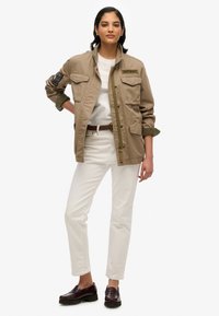 Khaki utilityjack met twee borstzakken, groene manchetknopen en "Superdry"-logo. Gecombineerd met witte broek en bordeauxrode instappers.