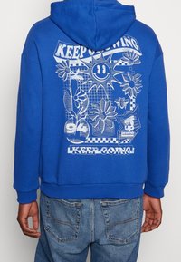 Sweat à capuche bleu avec un motif graphique comprenant des fleurs, un visage souriant et du texte au dos. Tissu en coton, capuche avec cordon de serrage, poignets côtelés.