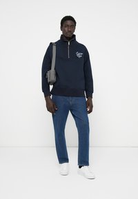 Navy fleece pullover with a quarter zipper, featuring a logo on the chest. Paired with blue denim jeans and white sneakers. Crossbody bag worn.