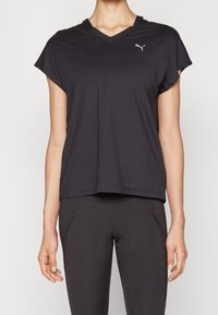 Kvinna klädd i svart V-ringad Puma T-shirt och svarta åtsittande tights, stående med avslappnade armar vid sidorna mot en enfärgad bakgrund.