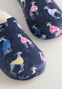 Marineblaue Hausschuhe mit einem Muster aus grauen Hunden, die bunte Pullover und Kleider tragen, auf einem hellen Untergrund.