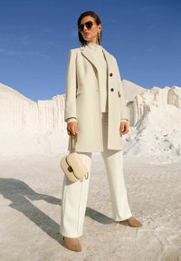 Vrouw in een beige jas, witte broek, beige enkellaarsjes, met een crèmekleurige handtas met gouden ketting, staande in een zandachtig witte omgeving onder een blauwe lucht.