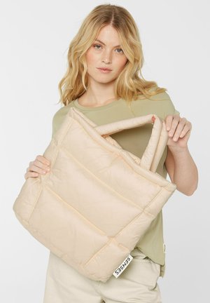 Padded beige tote bag with quilted design and dual top handles, featuring a label on one side. Model holds bag against a plain background.