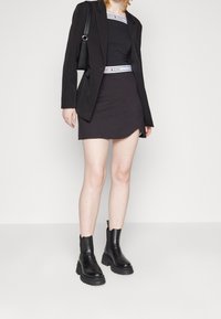 Musta bleiseri mustan, vartalonmyötäisen mekon päällä, jossa on kaartuva helma, yhdistettynä paksupohjaisiin mustiin nilkkurit; mekossa on brändätty joustava vyötärönauha.