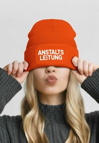 Orange knit beanie with a white patch reading "ANSTALTS LEITUNG." Model's hands hold the beanie down, partially covering the face. Gray sweater.