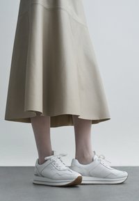 Zapatillas deportivas de cuero blanco con un acabado texturizado, que cuentan con cordones, suela de goma y un logo sutil, combinadas con una falda beige y fluida.