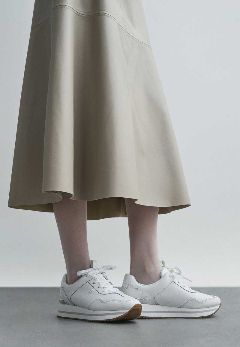 Zapatillas deportivas de cuero blanco con un acabado texturizado, que cuentan con cordones, suela de goma y un logo sutil, combinadas con una falda beige y fluida.