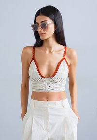 Crochet crop top in cream with red detailing, featuring a deep V-neck, adjustable straps, and a fitted design. Paired with cream high-waisted pants.