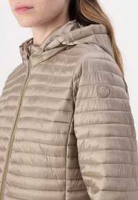 Beiger Puffer-Jacke mit horizontal gestepptem Muster, abnehmbarer Kapuze und goldfarbenem Reißverschluss. Verfügt über ein dezentes Logo am Oberarm.